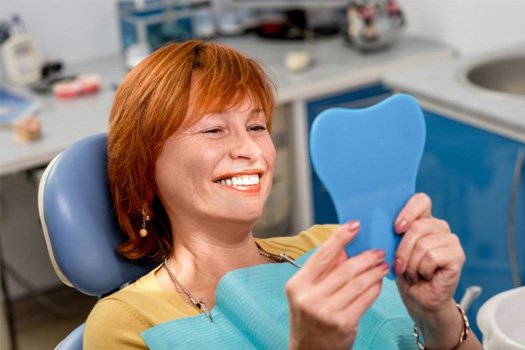 Woman in patient’s chair admiring her new smile in handheld mirror